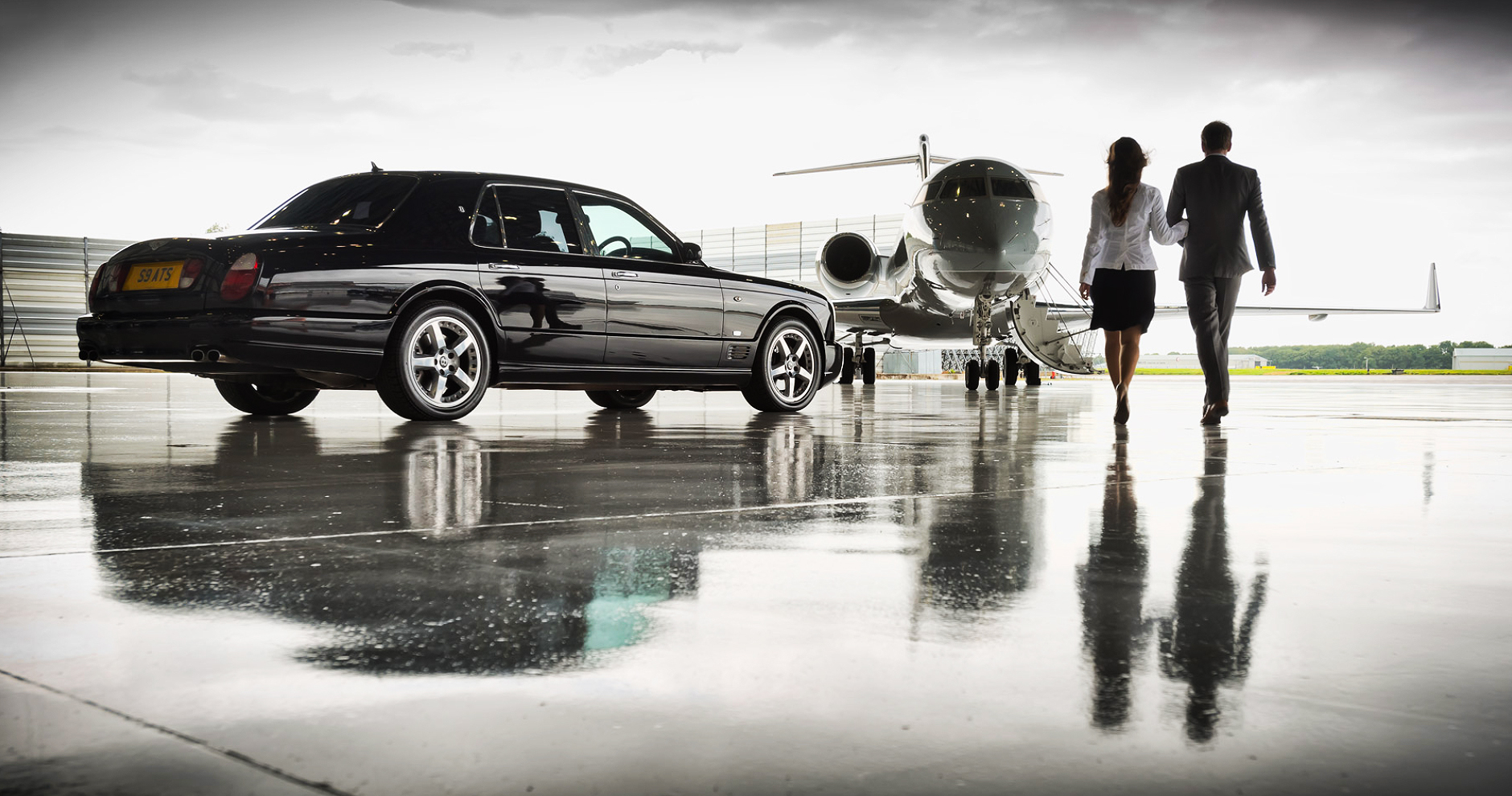 Executive couple walking toward a corporate jet from their Bentley on a wet airport ramp